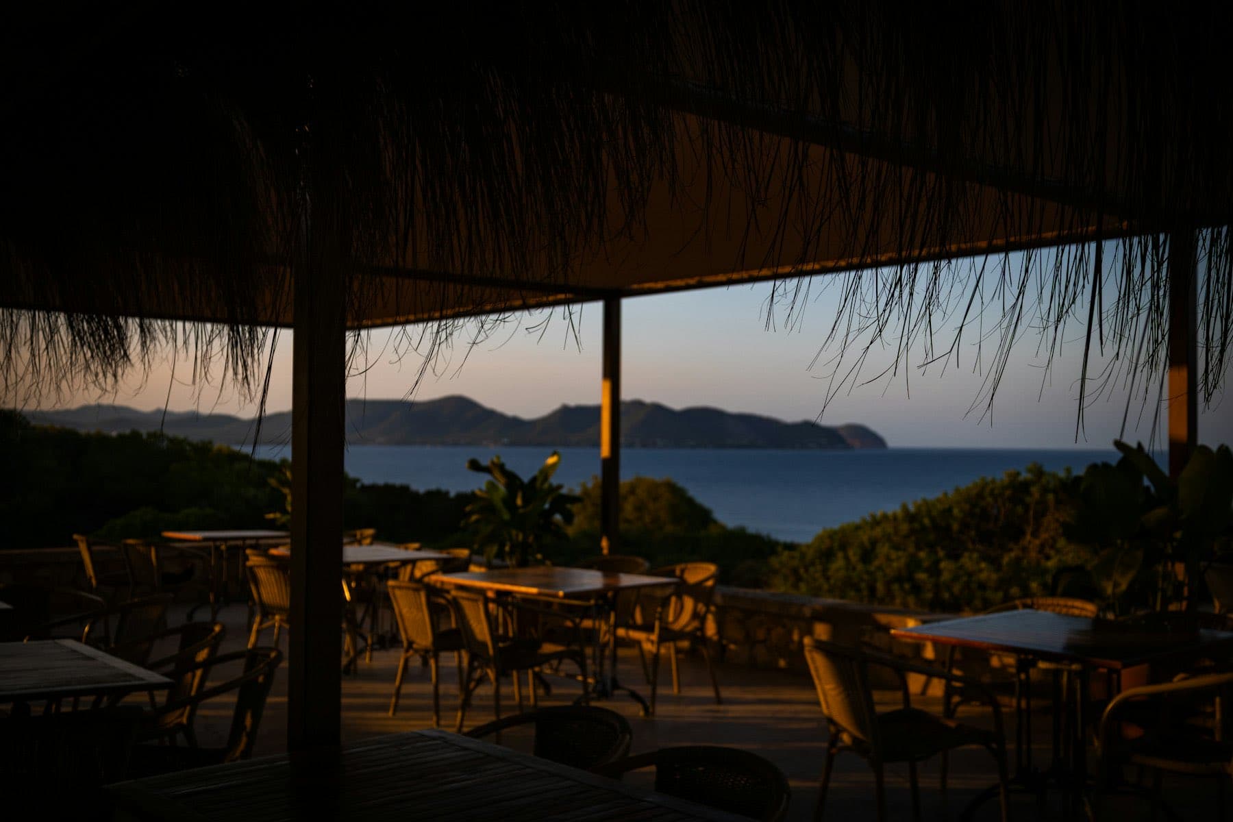Terraza bajo palapa con mesas mirando hacia la bahía al atardecer
